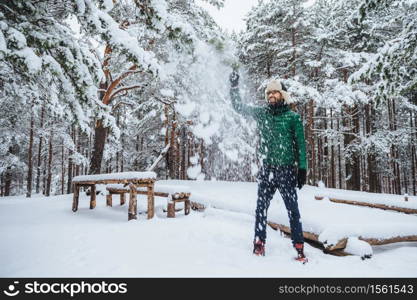 Outdoor shot of handsome bearded male dressed in warm clothes, has fun as throws snow in air, spends holidays in winter forest, expresses positiveness. Recreation and weather concept