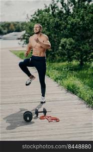 Outdoor shot of handsome fit muscular bearded man does high knee tap exercise, warms up before running, has cardio training, uses sport equipment, has strong body, demonstrates determination