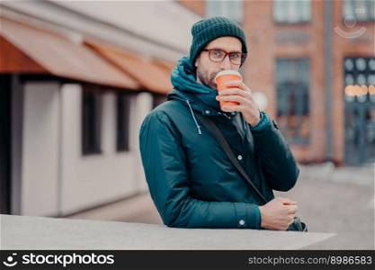 Outdoor shot of handsome hipster drinks takeawy coffee from disposable cup, dressed in headgear, jacket, wears spectacles, has srtoll outside, has straight look at camera. People and lifestyle