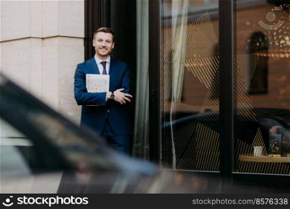 Outdoor shot of handsome male in formal clothes, stands crossed hands, holds journal, stands near cafe, waits for colleagues, has satisfied expression, gentle smile on face. People and lifestyle