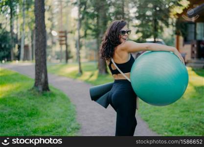 Outdoor shot of happy brunette woman carries fitball and fitness mat, wears sunglasses and sport clothes, poses in green park, being in good physical shape. Healthy lifestyle and hobby concept