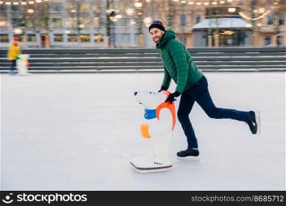 Outdoor shot of happy delightful man with thick beard and mustache looks gladfully into camera, demonstrates going skating on ice ring with skate aid, glad to go in for sport in such way
