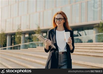 Outdoor shot of happy red haired businesswoman wears elegant formal clothes drinks takeaway coffee carries notebook and modern gadget necessary for work poses on stairs near office building.. Businesswoman wears elegant formal clothes drinks takeaway coffee carries notebook and modern gadget