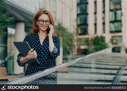 Outdoor shot of happy redhead woman in trendy clothes has telephone conversation walks on street poses near modern building holds laptop stands on stairs has cheerful expression. Lifestyle concept. Redhead woman in trendy clothes has telephone conversation walks on street near modern building