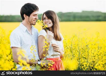 Outdoor shot of lovely couple stand close to each other, embrace and look in eyes, pose against yellow field background, enjoy calm atmosphere and harmony with nature. Love and people concept