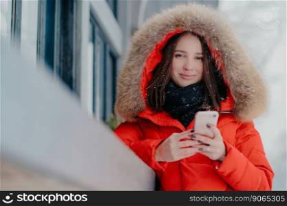 Outdoor shot of lovely dark haired Caucasian woman in winter street clothes, holds modern cell phone, reads received message, surfes social networks, enjoys spare time and snow, chilly weather