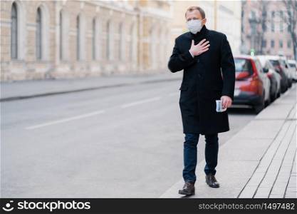 Outdoor shot of man goes to work, has problems with breathing, keeps hand on breast, wears medical mask, concerned with pandemic disease, poses outdoor. Viruses attack, health problem concept