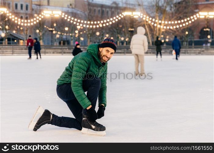 Outdoor shot of man in green coat, laces up skates, looks happily into camera, stands on wonderful ice ring decorated with garlands, has active lifestyle. People, leisure and winter concept.