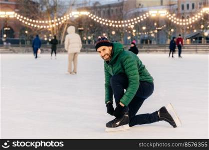 Outdoor shot of man in green coat, laces up skates, looks happily into camera, stands on wonderful ice ring decorated with garlands, has active lifestyle. People, leisure and winter concept.