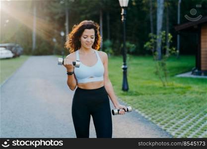Outdoor shot of motivated brunette young woman wears cropped bra and leggings, has slim perfect figure, raises dumbbells and trains biceps, has workout in open air. Fitness exercising concept