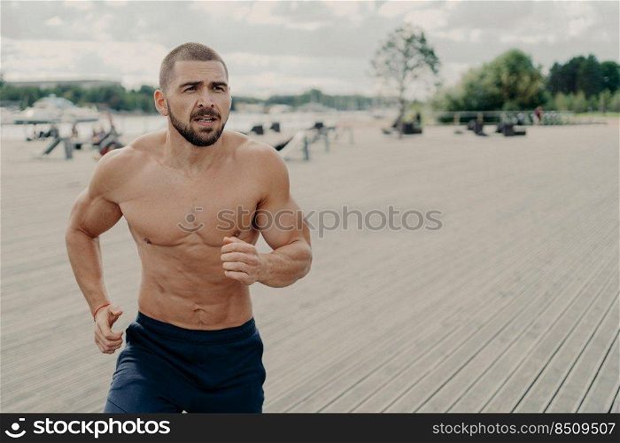 Outdoor shot of motivated shirtless muscular Caucasian man prepares for running marathon, goes jogging early in morning regularly, concentrated somewhere into distance. Sport and success concept