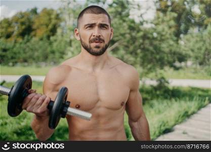 Outdoor shot of muscular bodybuilder with naked torso raises barbell and has strong biceps workout in open air looks thoughtfully into distance uses sport equipment. Handsome athlete with dumbbell