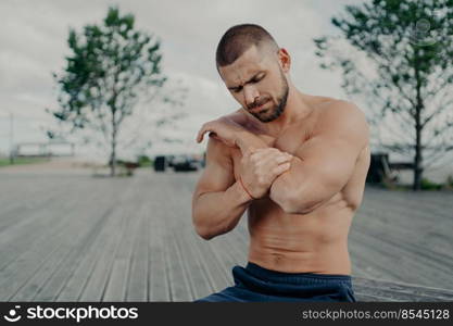 Outdoor shot of muscular guy injured elbow during physical exercises, feels strong pain and poses with naked torso. Health problems, medical, sickness and sport concept. Pain relief. Muscle injury