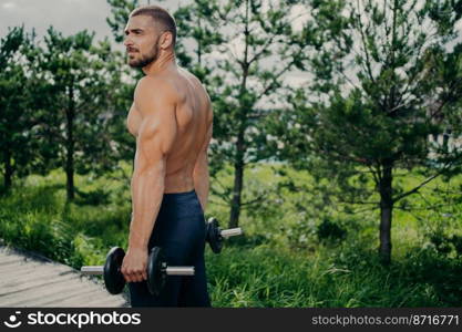 Outdoor shot of muscular strong man prepares for barbell workout, lifts weight and poses at street, gives motivation to everyone. Male weightlifter does biceps exercises, wears sportive clothes
