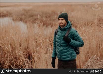 Outdoor shot of pensive male traveler dressed in warm clothes, being in good mood, has rucksack on back, walks in rural setting near field during autumn, enjoys calm atmosphere. Leisure concept