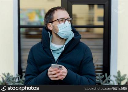 Outdoor shot of pensive man wears protective mask against coronavirus on street, holds mobile phone, reads news, cares about health, uses wireless internet. Health care. Covid-19. Virus spreading