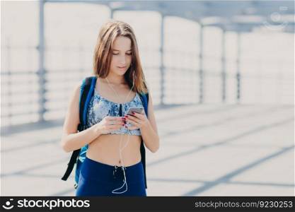 Outdoor shot of pleasant looking Caucasian female with slim body, carries rucksack, holds modern cell phone, listens radio online with earphones, reads notification, searches internet websites