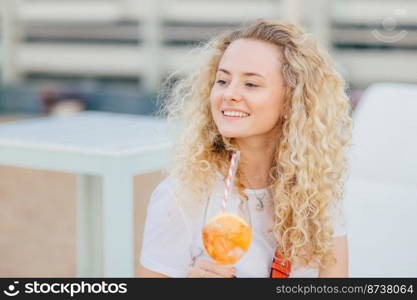 Outdoor shot of pleasant looking curly female looks happily aside, notices someone in distance, holds frech cold beverage, poses against beach background. People, drink and lifestyle concept