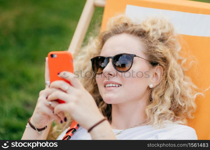 Outdoor shot of pleasant looking curly young female in sunglasses, holds modern orange smart phone, surfes social networks, connected to wireless internet, recreats in hammock during sunny day