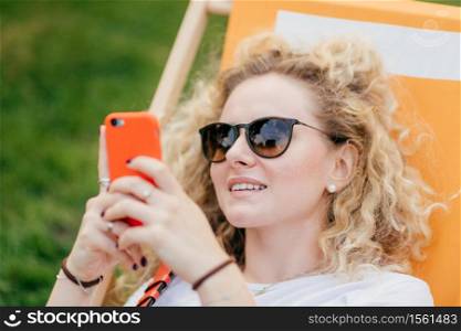 Outdoor shot of pleasant looking curly young female in sunglasses, holds modern orange smart phone, surfes social networks, connected to wireless internet, recreats in hammock during sunny day