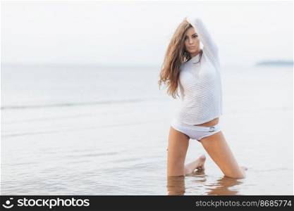 Outdoor shot of pleasant looking female model with slim perfect body shape, wears bikini, stands on knees at coastline, relaxes alone during summer holidays. Fit young woman with attractive look