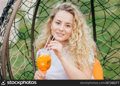 Outdoor shot of pleasant looking female with curly light hair, drinks fresh cold drink, rests in hanging chair, breathes fresh air, has summer party with friends. People and lifestyle concept.