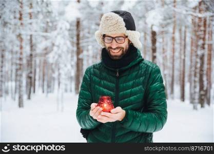 Outdoor shot of pleasant looking male holds candle in hands, warms hands, looks happily on it as stands in winter forest covered with snow. Positive young Caucasian man enjoys beautiful nature