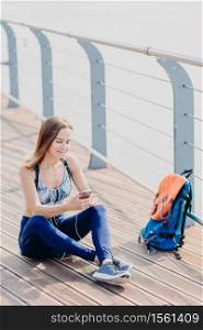 Outdoor shot of pleasant looking relaxed woman sits crossed legs, looks positively at smart phone, listens music with earphones enjoys high speed internet. Female traveller takes break after long walk