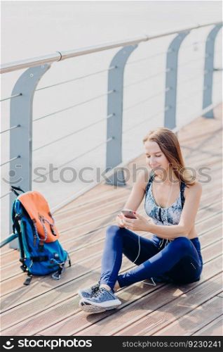 Outdoor shot of pleasant looking relaxed woman sits crossed legs, looks positively at smart phone, listens music with earphones enjoys high speed internet. Female traveller takes break after long walk