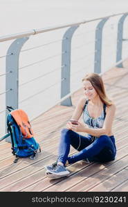 Outdoor shot of pleasant looking relaxed woman sits crossed legs, looks positively at smart phone, listens music with earphones enjoys high speed internet. Female traveller takes break after long walk