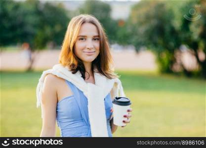 Outdoor shot of pleasant looking young European female with dark hair, holds takeaway coffee, spends free time in park, wears casual t shirt and sweater on shoulders, looks directly at camera