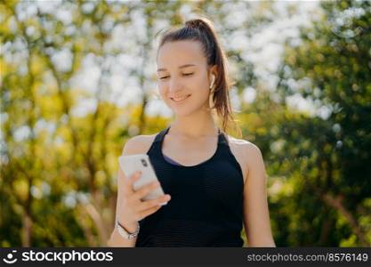 Outdoor shot of pleased sporty woman focused at smartphone checks how many calories she burnt after jogging exercise uses special app listens music in earphones has pony tail wears black t shirt
