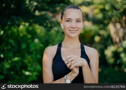 Outdoor shot of pretty dark haired woman keeps hands together being in good physical shape as goes in for sport regularly wears sportsclothes listens music while having workout at green park