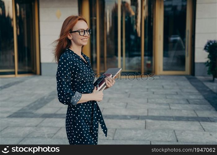 Outdoor shot of prosperous skilled redhead businesswoman stands sideways to camera has cheerful expression carries modern gadgets necessary for work notepad wears polka dot dress and spectacles. Skilled redhead businesswoman stands sideways has cheerful expression carries modern gadgets