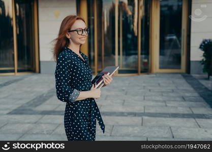 Outdoor shot of prosperous skilled redhead businesswoman stands sideways to camera has cheerful expression carries modern gadgets necessary for work notepad wears polka dot dress and spectacles. Skilled redhead businesswoman stands sideways has cheerful expression carries modern gadgets