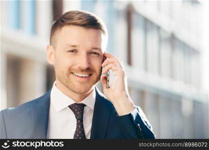 Outdoor shot of prosperous successful unshaven businessman wears formal clothes, has pleasant talk with business partner via cell phone, discuss future meeting, poses against blurred background