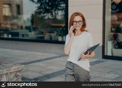 Outdoor shot of red haired woman has telephone conversation discusses coming meeting with friend carries tablet notebook looks away into distance wears casual clothes satisfied with good connection. Woman has telephone conversation discusses coming meeting with friend carries tablet notebook