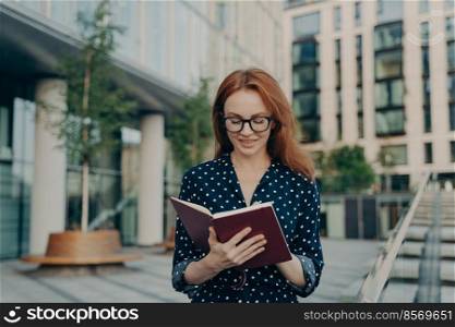 Outdoor shot of red haired woman makes notes in notepad makes plannings for working day wears spectacles polka dot dress poses near office building writes down list to do. Busy businesswoman. Outdoor shot of red haired woman makes notes in notepad makes plannings for working day