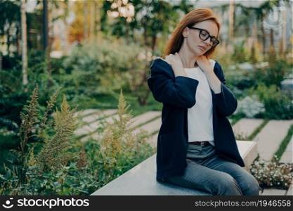 Outdoor shot of redhead woman has shoulder pain body muscles problems wears transparent glasses and formal clothes focused down poses outdoor in park during daytime against blurred background. Outdoor shot of redhead woman has shoulder pain poses outdoor in park during daytime