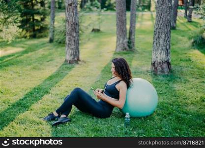 Outdoor shot of relaxed brunette woman sits near fitness ball, uses modern mobile phone, listens music via earphones, poses on green grass with bottle of water, wears sports clothes and sneakers