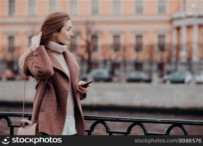 Outdoor shot of relaxed young female looks aside with thoughtful expression, holds modern cell phone, waits for call, dressed in white sweater and coat, carries bag, stand on bridge in big city
