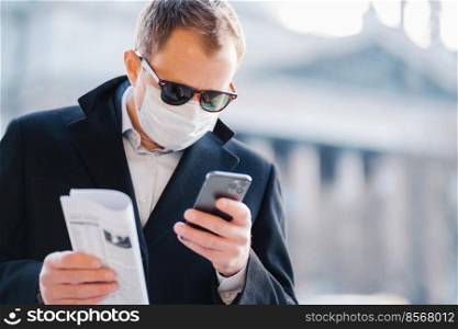 Outdoor shot of serious man manager focused in cellular display, reads news online, holds newspaper, wears medical mask to protect from coronavirus, infection with influenza virus, poses on street