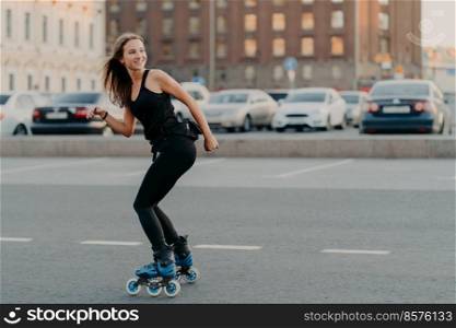 Outdoor shot of slim cheerful woman dressed in active wear looks away leads healthy lifestyle rides on rollers poses on road in urban place feels energized. Rollerblading as hobby. Recreation