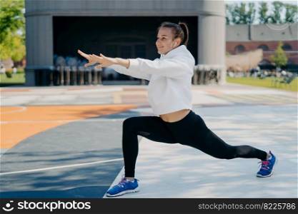 Outdoor shot of slim sporty woman involved in recreation activities stretches arms legs looks into distance wears white hoodie leggings and sportshoes stetches body before run leads healthy lifestyle
