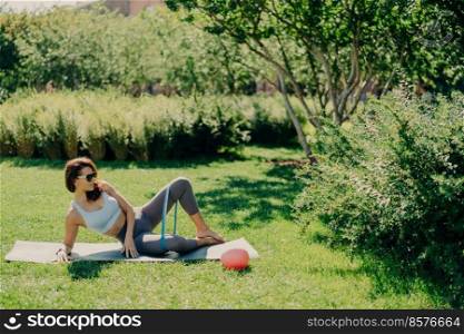 Outdoor shot of slim sporty young woman in activewear practices exercises for legs with elastic band lying on karemat at green grass enjoys fresh air keeps fit. People fitness aerobics concept