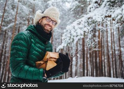 Outdoor shot of smiling glad male with beard and mustache wears spectacles, anork and warm hat, holds firewood, stands against trees covered with snow, enjoys fresh frosty air in forest