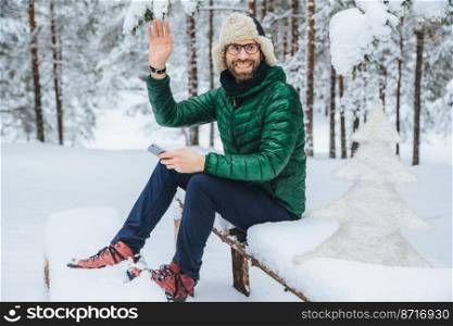 Outdoor shot of smiling male wears spectacles dressed in warm clothes, waves with hand as notices someone, holds smartphone, sits on wooden bench in winter forest, gestures actively