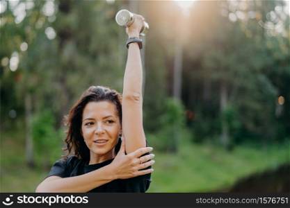 Outdoor shot of sporty European woman has dark hair raises arm with dumbbell, trains muscles and looks somewhere into distance, poses over green nature background. Female with sport equipment