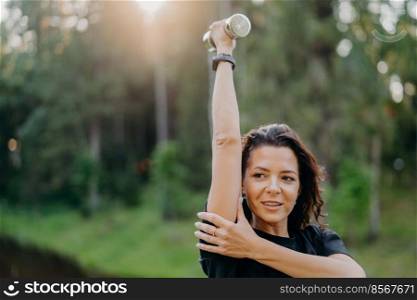 Outdoor shot of sporty European woman has dark hair raises arm with dumbbell, trains muscles and looks somewhere into distance, poses over green nature background. Female with sport equipment