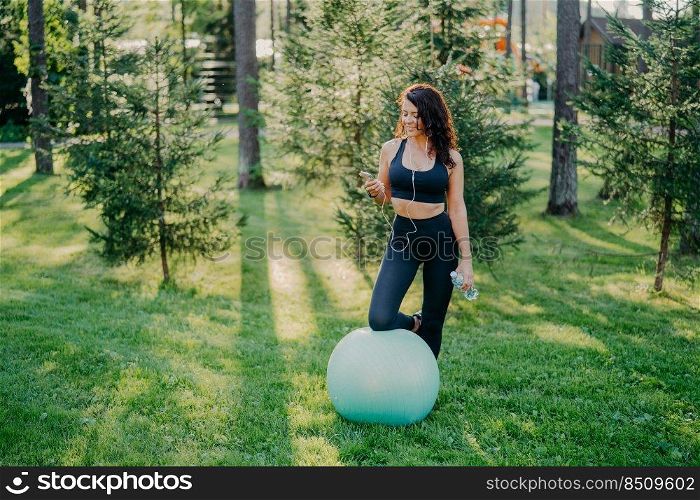 Outdoor shot of sporty young woman holds bottle of fresh water, listens music in earphones, holds modern smartphone, leans knee on fitness ball, enjoys favorite playlist, poses in green forest